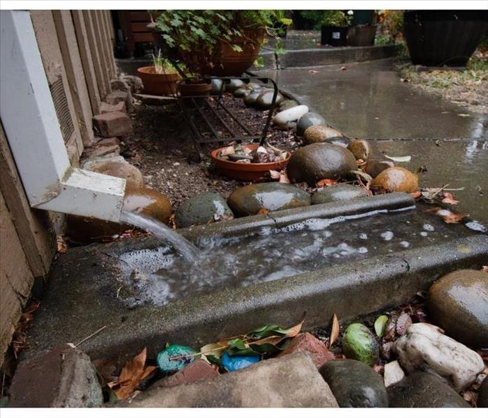 A downspout on a house, water pouring onto a concrete splash block, and over a sidewalk path during heavy rain